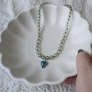 May include: A beaded necklace with light green beads and black spacers. A teal heart pendant hangs from the center. The necklace is displayed on a white, scalloped dish.