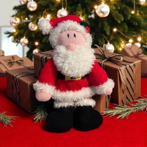 May include: A knitted Santa Claus doll in a red suit, white beard, and black boots. The doll has a pink face and a red hat with white trim. It is on a red surface with wrapped gifts behind it, and a Christmas tree in the background.