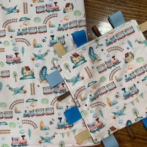 May include: A white baby blanket with a train and airplane pattern. The blanket has a matching tag with blue, brown, and beige fabric ties.