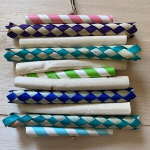 May include: A vibrant bird toy with a mix of textures and colours. It features woven sections in blue, purple, and teal, alongside white and striped paper rolls in pink, green, and blue. A metal clip is attached for hanging.