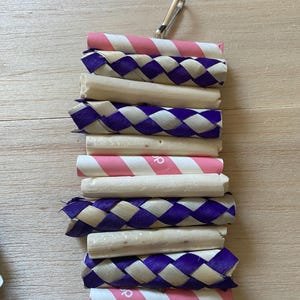 May include: A bird toy composed of woven purple and white material, natural-coloured wood, and pink and white striped paper tubes. A silver metal clip is attached to the top for hanging.