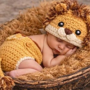 Op de afbeelding: Een slapende baby draagt een gehaakt leeuwenkostuum, inclusief een hoed en een korte broek, en rust in een gevlochten mand gevuld met bruine vezels. De leeuwenhoed heeft een bruine manen, oren en een gezicht met zwarte ogen en een bruine neus.