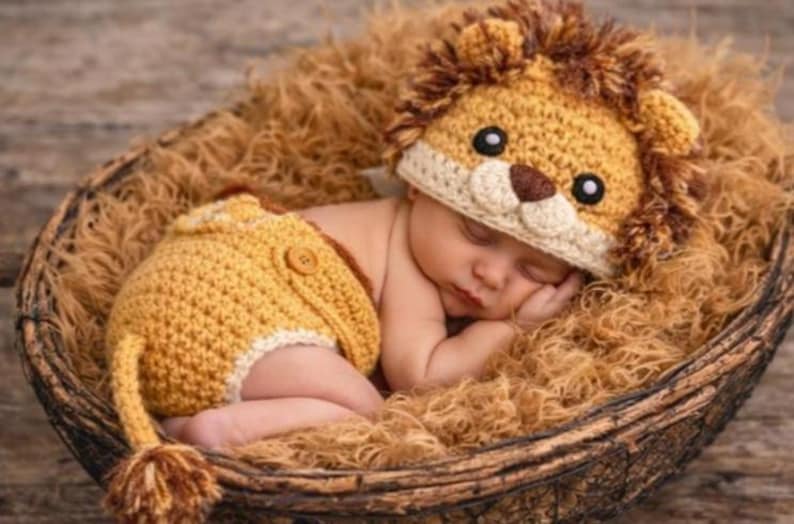Op de afbeelding: Een slapende baby draagt een gehaakt leeuwenkostuum, inclusief een hoed en een korte broek, en rust in een gevlochten mand gevuld met bruine vezels. De leeuwenhoed heeft een bruine manen, oren en een gezicht met zwarte ogen en een bruine neus.