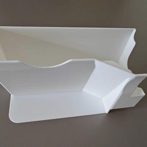 May include: A white, angular, 3D-printed organizer with a unique design. It features a main compartment with a curved edge and a smaller, angled section. The overall shape is geometric and modern, suitable for desk organization.