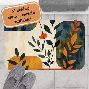 May include: A rectangular bath mat featuring an abstract botanical design with dark green and orange leaves, a single red flower, and geometric shapes in cream, mustard yellow, and dark blue. The mat has rounded corners and a smooth surface texture.