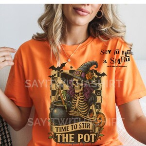 May include: Orange t-shirt with a Halloween-themed graphic. The design features a skeleton witch stirring a cauldron, with text that reads "Time to Stir the Pot." The shirt also includes the text "Say it with a Shirt."
