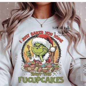 May include: White sweatshirt featuring a Grinch character wearing a Santa hat, holding a spoon, and surrounded by cupcakes. The text reads "I JUST BAKED YOU SOME FUCUPCAKES." A white mug with a snowflake design is also visible.