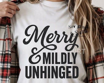 May include: White t-shirt with the text "Merry & Mildly Unhinged" in a black, cursive font. The shirt also has the text "Say it with a Shirt" in a smaller font. The person is wearing a red and black plaid shirt over the white t-shirt.