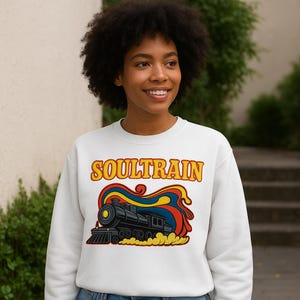 May include: White sweatshirt featuring the text "SOULTRAIN" in yellow and orange lettering above a colorful graphic of a black train with red, blue, and yellow accents. The sweatshirt is a crew neck style.