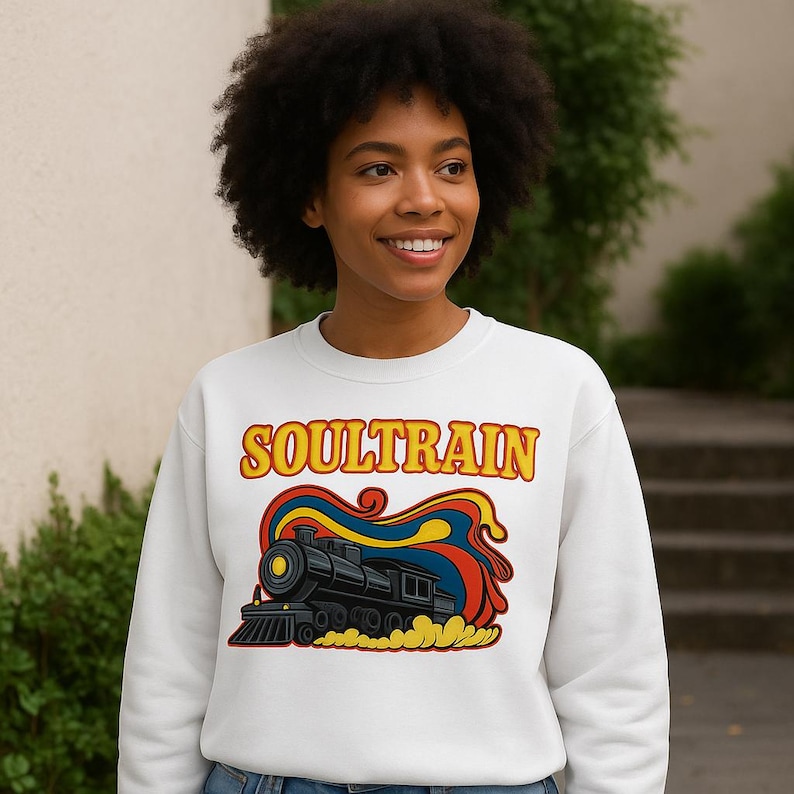 May include: White sweatshirt featuring the text "SOULTRAIN" in yellow and orange lettering above a colorful graphic of a black train with red, blue, and yellow accents. The sweatshirt is a crew neck style.