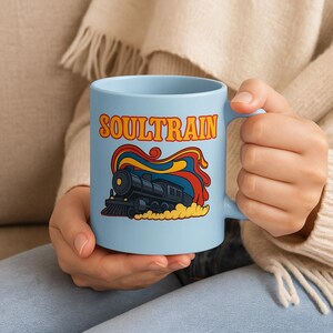 May include: A light blue ceramic mug featuring the word "SOULTRAIN" in orange and yellow retro font. Below the text is a black train illustration with red, yellow, and blue accents. The mug is held by a person.