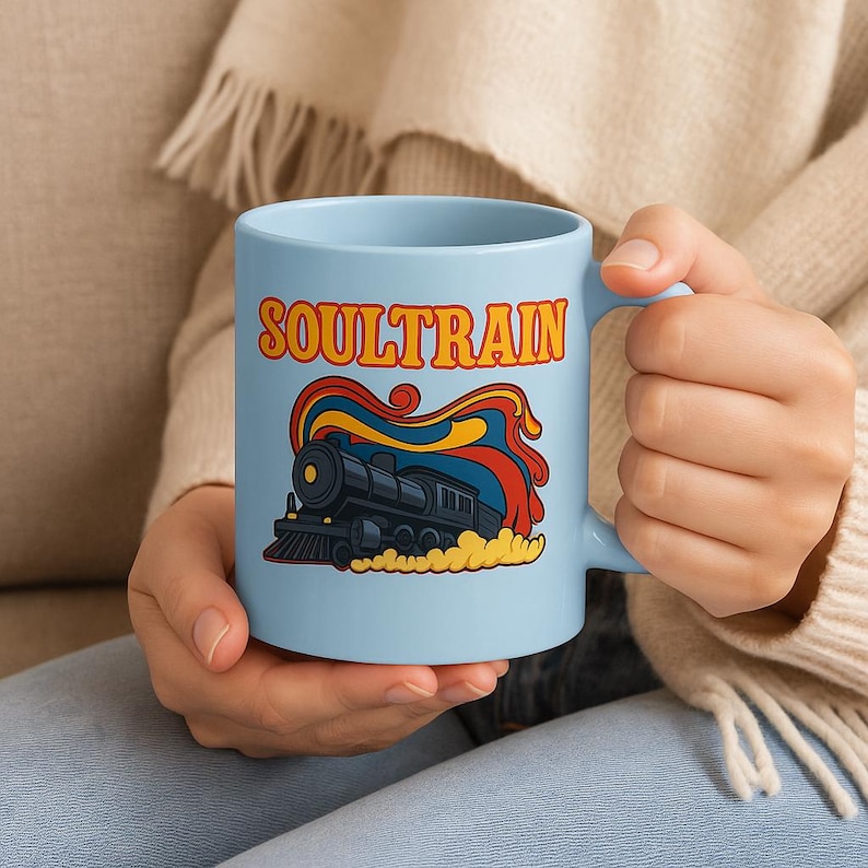 May include: A light blue ceramic mug featuring the word "SOULTRAIN" in orange and yellow retro font. Below the text is a black train illustration with red, yellow, and blue accents. The mug is held by a person.