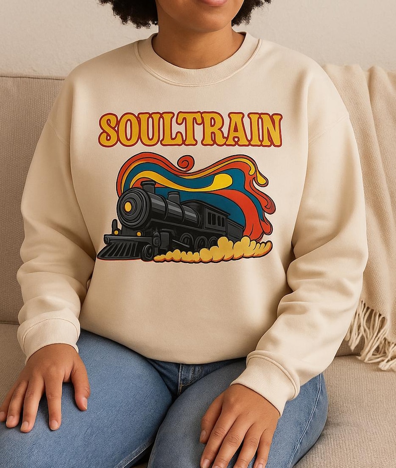 May include: Cream-colored sweatshirt featuring a retro graphic of a black train with colorful smoke and the word "SOULTRAIN" in orange letters. The sweatshirt has long sleeves and a crew neck.