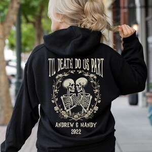May include: Black hoodie with the text "TIL DEATH DO US PART" above a graphic of two skeletons embracing within a floral wreath. Below the graphic are the names "ANDREW & MANDY" and the year "2022".