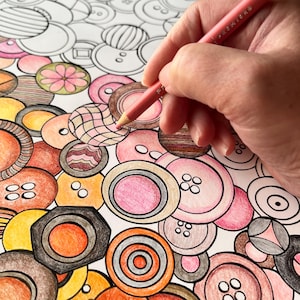 May include: A colouring page featuring a pattern of various circular buttons in shades of pink, orange, yellow, and brown. A hand holds a pink coloured pencil, actively filling in a button with a grid design on the white paper surface.