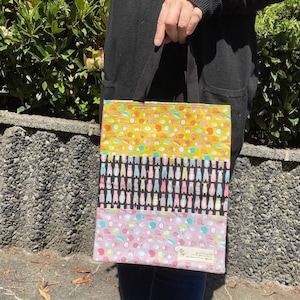 May include: A tote bag with a multi-coloured pattern. The bag has three panels, each with a different pattern. The top panel is yellow with a small, repeated pattern of animals. The middle panel is black with a repeated pattern of animals in various colours. The bottom panel is light purple with a repeated pattern of animals. The bag has a black handle.