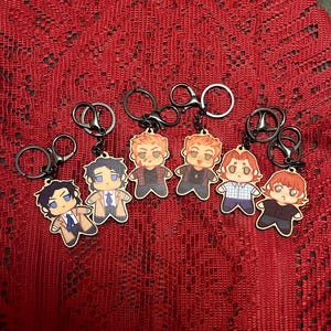 May include: Six keyrings with cartoon character charms. Each charm is a different character, with various outfits and hairstyles. The keyrings have black metal rings. The charms appear to be made of wood or a similar material.