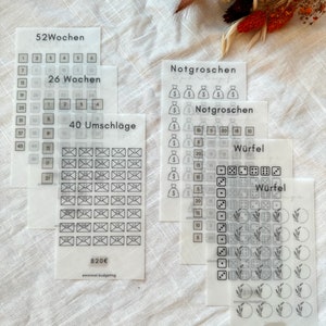 Könnte beinhalten: Vier transparente Blätter mit schwarzen Umrissen verschiedener Budgetierungstools. Das erste Blatt hat ein Raster mit den Zahlen 1-52 und dem Text "52 Wochen" und "26 Wochen". Das zweite Blatt hat ein Raster mit 40 Umschlägen und dem Text "40 Umschläge". Das dritte Blatt hat ein Raster mit Geldbeuteln und dem Text "Notgroschen" und "Notgroschen". Das vierte Blatt hat ein Raster mit Würfeln und dem Text "Würfel" und "Würfel".