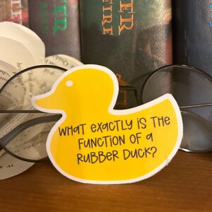 May include: A yellow rubber duck sticker with the text "WHAT EXACTLY IS THE FUNCTION OF A RUBBER DUCK?" on a wooden surface.