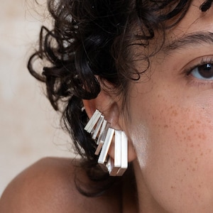 Può includere: Orecchino a cuffia in argento con un design geometrico. Il bracciale è composto da più pezzi rettangolari impilati uno sopra l'altro.