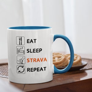 May include: White ceramic mug with a blue interior and handle. The mug features the words "EAT SLEEP STRAVA REPEAT" in black text and orange text. The mug is on a wooden table.
