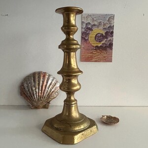 Könnte beinhalten: Ein goldfarbener Kerzenhalter mit quadratischem Sockel und dekorativem, mehrstufigem Design. Eine Muschel und ein kleines Gemälde mit einem Halbmond sind ebenfalls sichtbar.