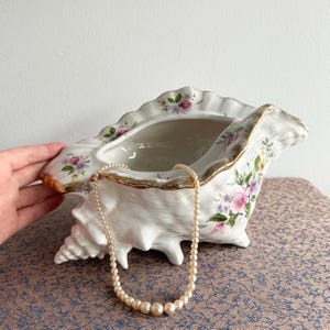 May include: A white ceramic seashell-shaped decorative bowl with gold trim and floral accents. A cream-colored pearl necklace rests inside the bowl. The bowl is on a patterned surface.