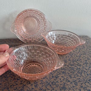 Könnte beinhalten: Drei rosa Glasschalen mit Griffen. Die Schalen haben ein gewebtes Muster und sind aus klarem Glas mit einer rosa Tönung.