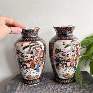 May include: Two porcelain vases with a blue and white floral design. The vases feature a scene of people in traditional Japanese clothing. The vases are decorated with gold accents.