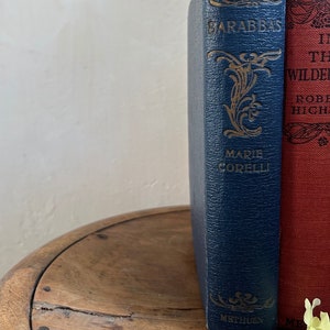 May include: Two vintage hardcover books with gold lettering on the spines. The book on the left is titled "Barabbas" by Marie Corelli and is bound in dark blue. The book on the right is titled "In the Wilderness" and is bound in red.