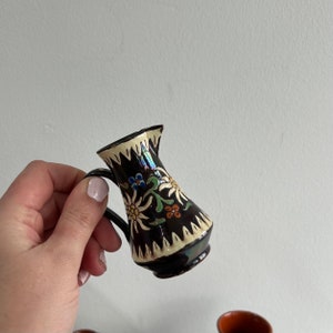 May include: A small brown ceramic jug with a white handle and floral design. The jug has a dark brown glaze with white and yellow flowers and green leaves. The jug is decorated with a white and black geometric pattern around the rim.