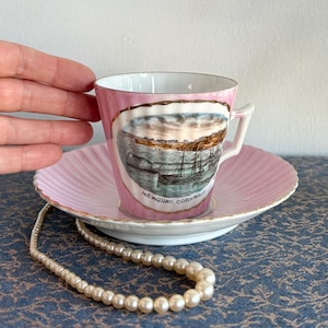 May include: A pink teacup and saucer with a gold rim. The teacup features a scenic image of Newquay, Cornwall, with a sailboat in the harbor.