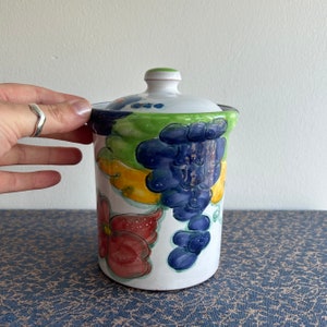 Peut inclure: Pot en céramique blanche avec un couvercle, décoré d'un motif peint à la main de raisins, de feuilles et de fleurs dans des tons de rouge, jaune, vert et bleu.