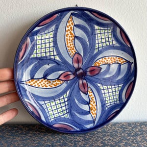May include: A blue and white ceramic plate with a floral design. The plate features a central flower with pink petals and a dark blue centre. The flower is surrounded by swirling blue and white lines and orange dots.