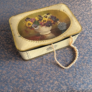 May include: A vintage tin box with a floral design featuring a bouquet of pansies in a white vase. The box is closed and a string of pearls is draped over the top.