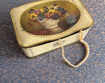 May include: A vintage tin box with a floral design featuring a bouquet of pansies in a white vase. The box is closed and a string of pearls is draped over the top.