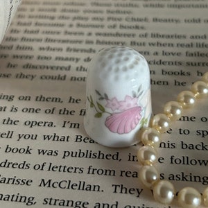 May include: A white porcelain thimble with a floral design and a pink seashell. The thimble is sitting on a book with a pearl necklace next to it.