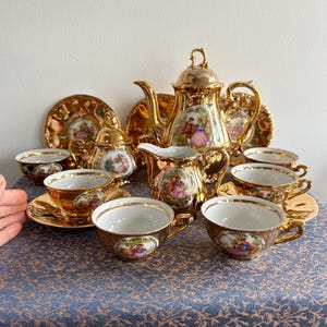 May include: A complete gold tea set including a teapot, creamer, sugar bowl, saucers, and cups. Each item has a painted scene of a couple in a garden. The set is displayed on a blue tablecloth.