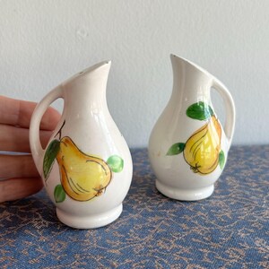 May include: Two white ceramic salt and pepper shakers. Each shaker is decorated with a yellow pear and green leaves. The shakers have a handle and a spout. They are set against a blue patterned surface.