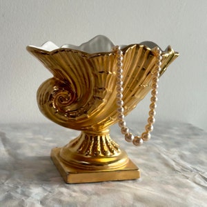 May include: A gold-plated ceramic seashell bowl with a white interior. The bowl is on a pedestal base and has a pearl necklace draped over it.