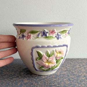 May include: A white ceramic planter with a purple rim and floral designs. The planter features pink, purple, and green flowers with a white background.