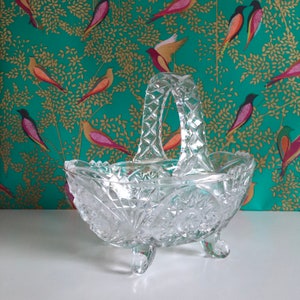 May include: A clear glass serving bowl with a decorative cut-glass pattern. The bowl has a handle and three legs. The bowl is on a white surface.