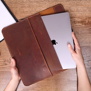 Peut inclure: Une pochette pour ordinateur portable en cuir marron avec une fermeture à rabat. La pochette est ouverte et un ordinateur portable argenté est partiellement visible à l'intérieur.