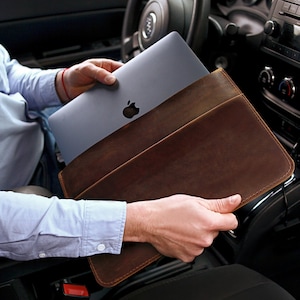 May include: A brown leather laptop sleeve with a laptop computer inside. The sleeve has a flap that closes with a magnetic snap. The laptop is a silver color with a white Apple logo.
