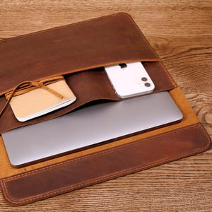 Peut inclure: Une pochette pour ordinateur portable en cuir marron avec une poche intégrée pour un téléphone et une plus petite poche pour un carnet. La pochette est ouverte et un ordinateur portable, un téléphone et un carnet sont à l'intérieur.