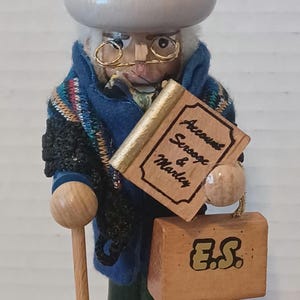May include: Wooden Scrooge figurine wearing a blue coat, top hat, and glasses. He holds a book labeled "Account Scrooge & Marley" and a wooden box with "E.S." on it. A cane rests at his side. The base reads "Scrooge".