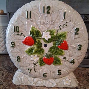 May include: A round ceramic quartz clock featuring a raised relief pattern of white leaves. The center displays three red strawberries with green leaves and small white flowers. Dark green numbers mark the hours, and the word QUARTZ is printed.