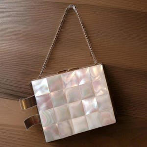 Puede incluir: Un bolso de mano cuadrado blanco con una correa de cadena. El bolso de mano tiene un patrón de cuadrícula de baldosas cuadradas con un brillo nacarado. Se ven detalles metálicos dorados en los lados.