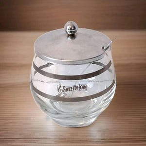 May include: A clear glass sugar bowl with a silver lid and a small spoon. The bowl has two horizontal silver bands and the text "Sweet'N Low" printed on the front. The lid has a small, round knob.