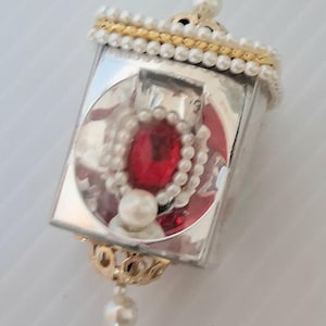May include: A decorative ornament featuring a square, mirrored frame with a large, oval red gem surrounded by pearls. The frame is accented with gold trim and a strand of pearls. A pearl strand hangs below.